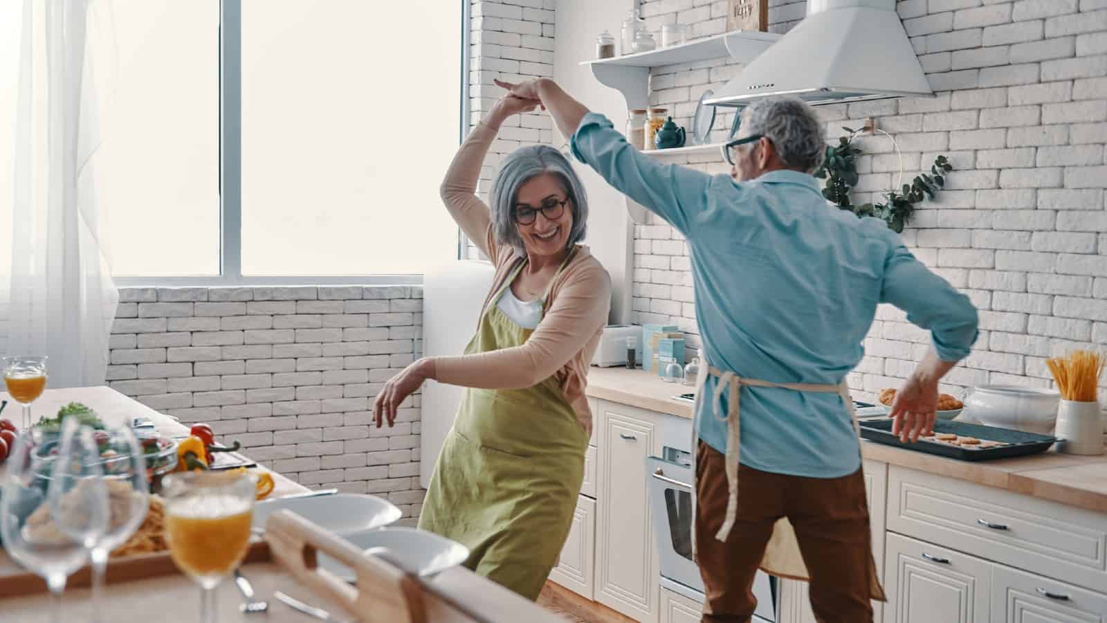 Playful senior couple in aprons dancing and smiling while preparing healthy dinner at home