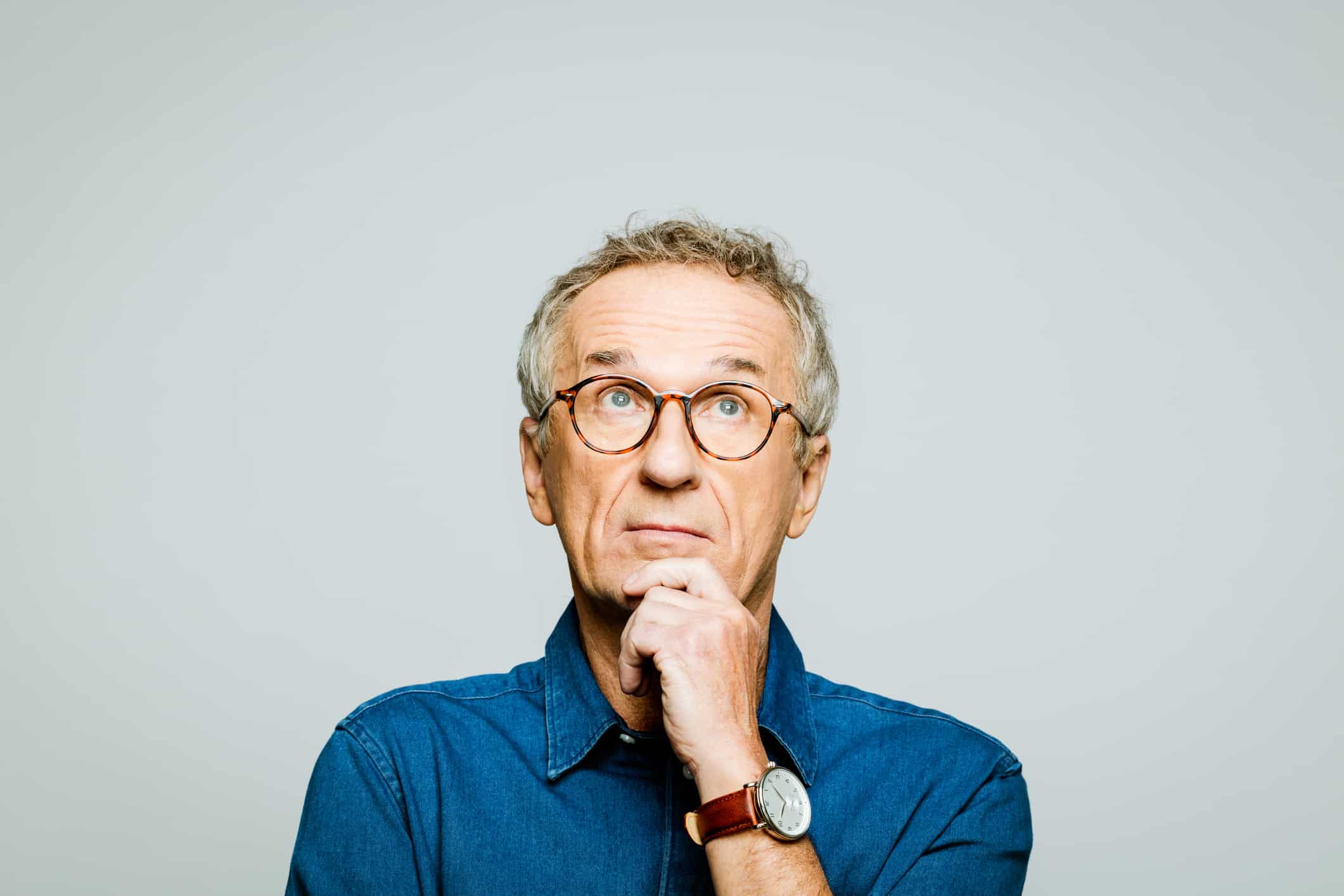 Portrait of elderly man wearing white denim shirt and glasses looking up with hand on chin. Thoughtful senior entrepreneur, studio shot against grey background.