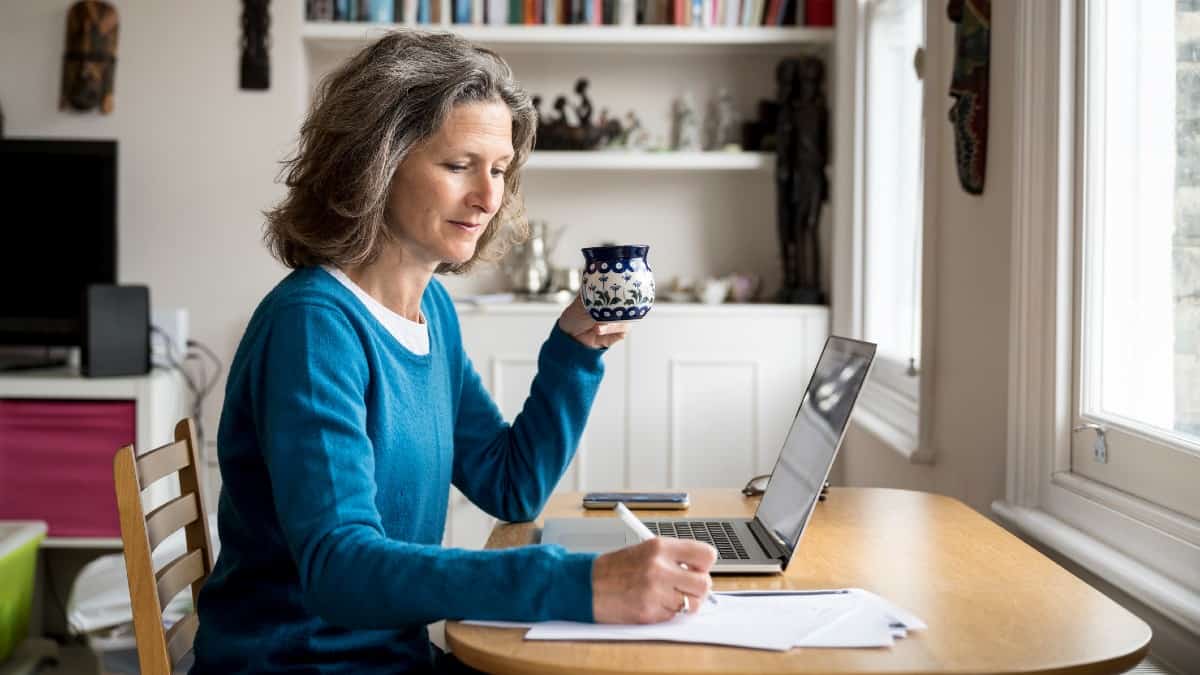 Mature Caucasian woman sat at a table with coffee and laptop while making notes on paper