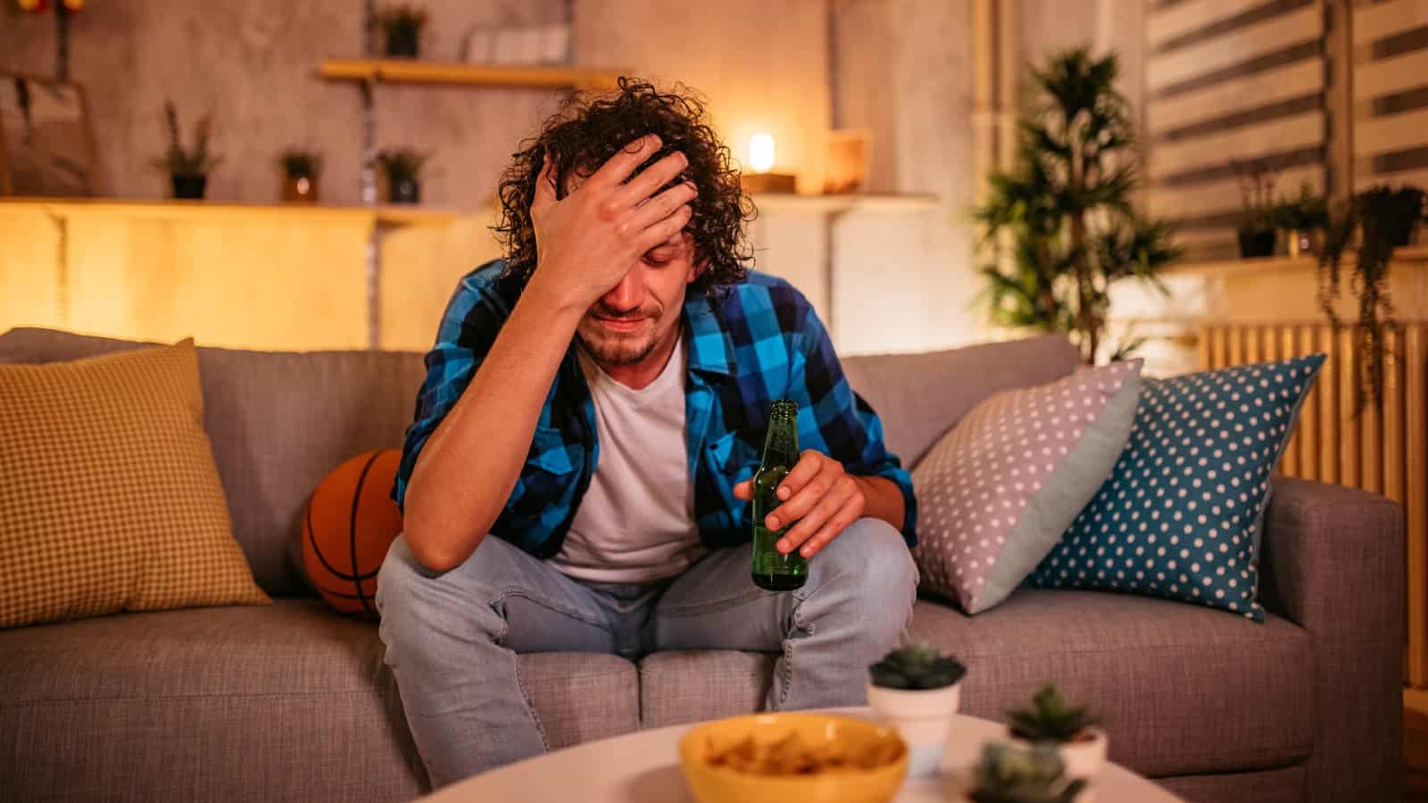 Frustrated young white male looking disconsolate while sat on his sofa holding a beer