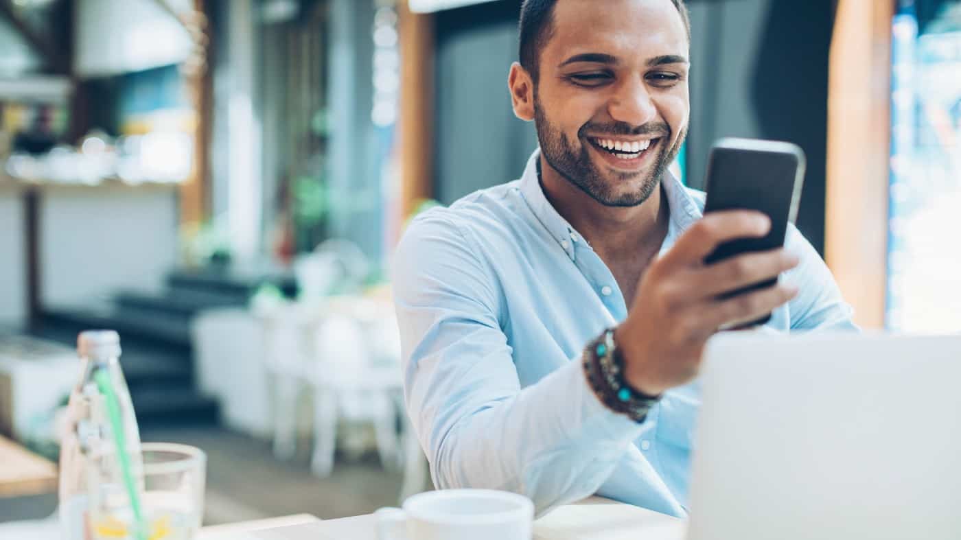 Smiling young man sitting in cafe and checking messages, with his laptop in front of him.