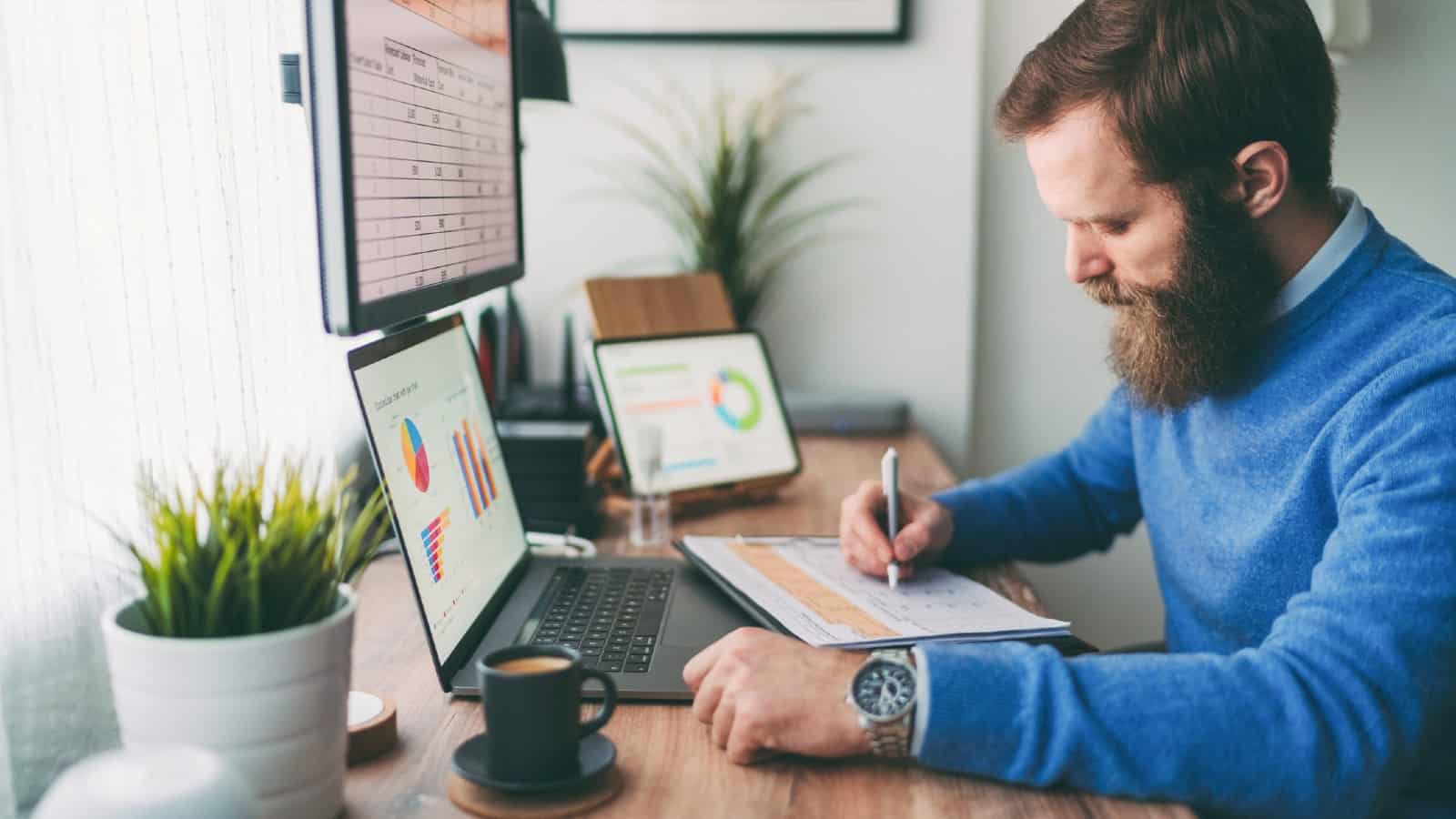 Bearded man writing on notepad in front of computer