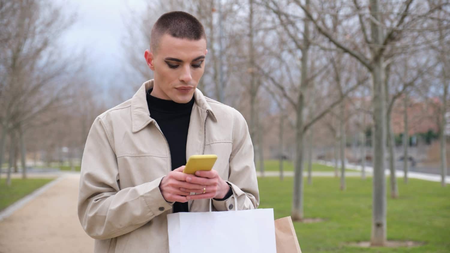 Handsome young non-binary androgynous guy, wearing make up, chatting on his smartphone, carrying shopping bags.