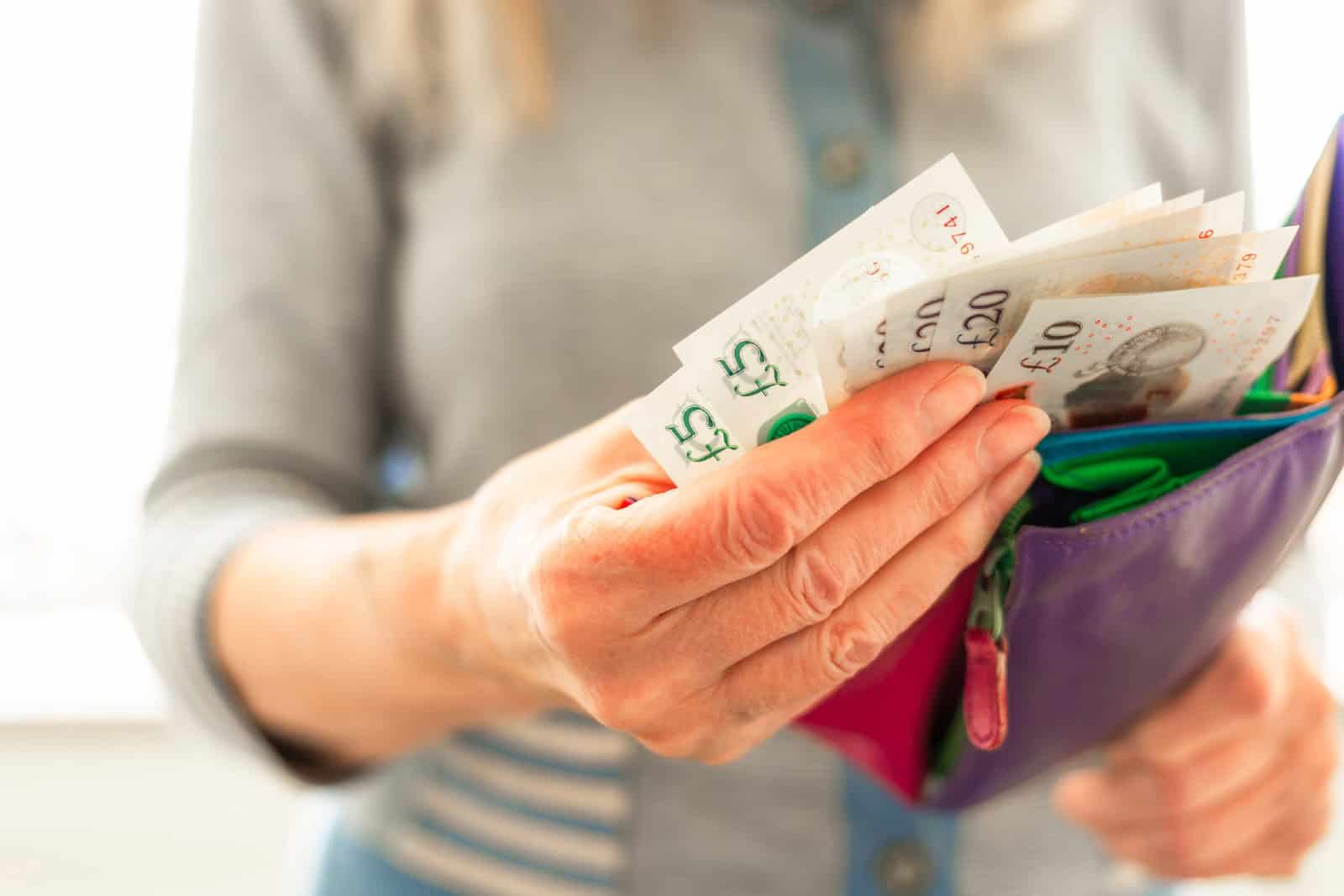 Close-up image depicting a woman in her 70s taking British bank notes from her colourful leather wallet.
