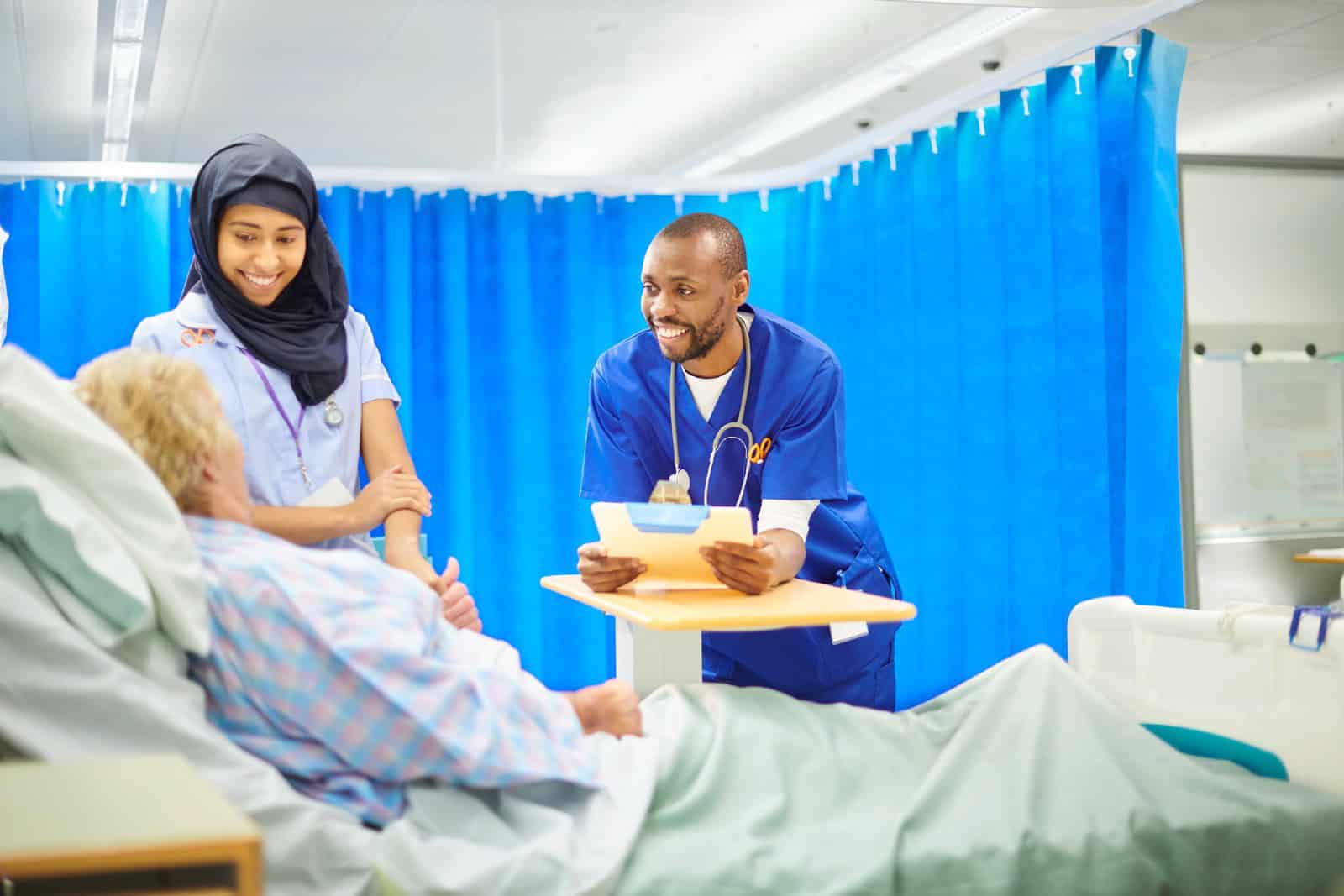 A black male doctor chats to a senior patient on the hospital ward ,with a young female nurse wearing a hijab attending to a dressing