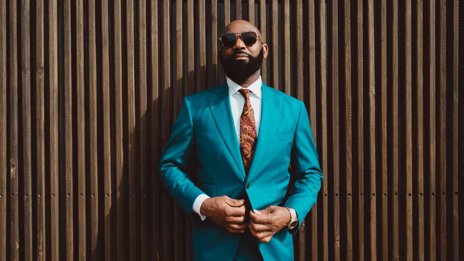 A handsome mature bald bearded black man in a sunglasses and a fashionable blue or teal costume with a tie is standing in front of a wall made of striped wooden timbers and fastening a suit button