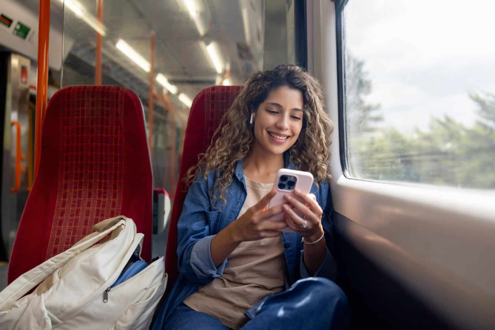 Happy woman commuting on a train and checking her mobile phone while using headphones